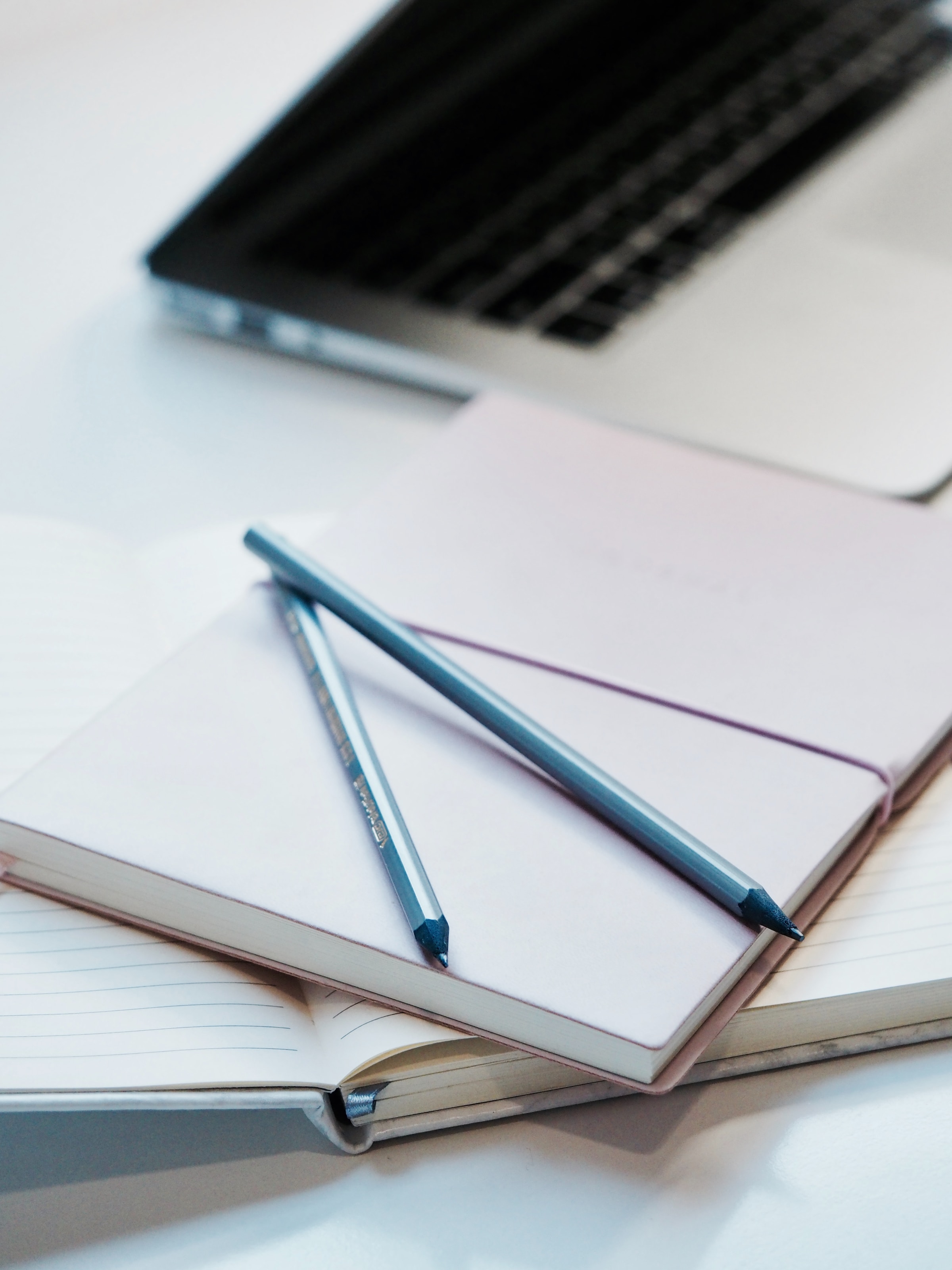 dos libretas sobre mesa blanca, con dos lápices y portátil de fondo