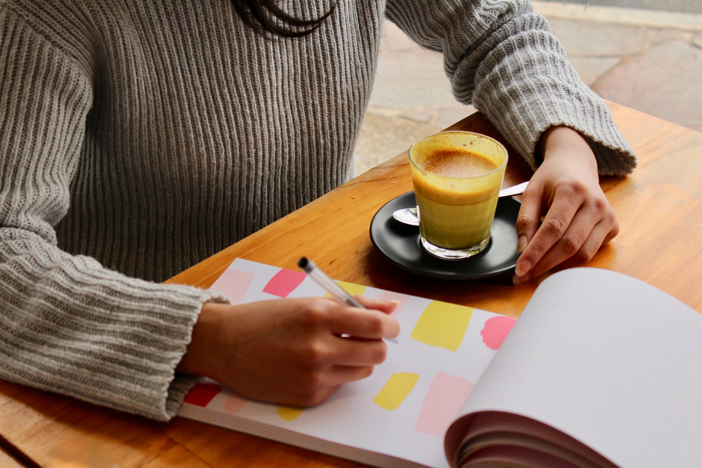 imagen de torso y brazos de una mujer escribiendo sobre un cuaderno con trazas de colores y una taza de café a la izquierda