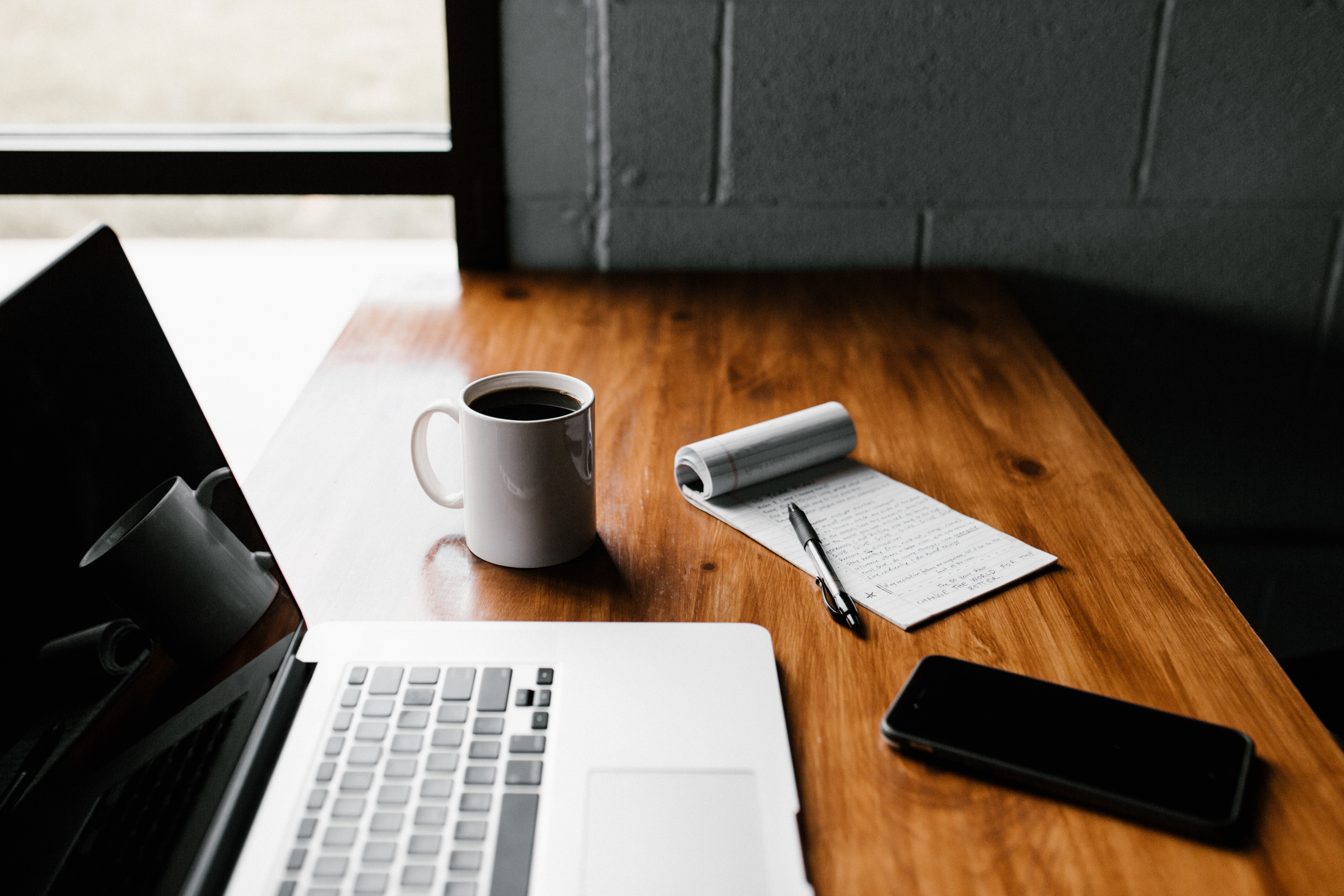 Imagen de una mesa de trabajo con ordenador portátil, teléfono móvil, taza de café y libreta
