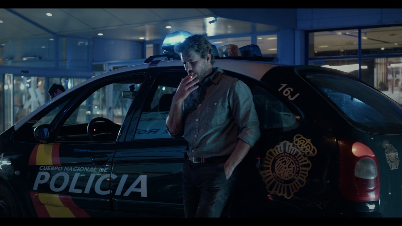 Fotograma de la película Delfines de plata. Rodolfo Sancho fumando apoyado sobre un coche de poliía nacional