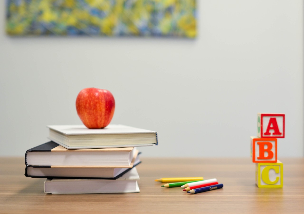 Imagen de una mesa con libros apilados con manzana encima y unos lapiceros de colores al lado. También a la derecha vemos unos cobos de colores apilados con las letras A,B,C